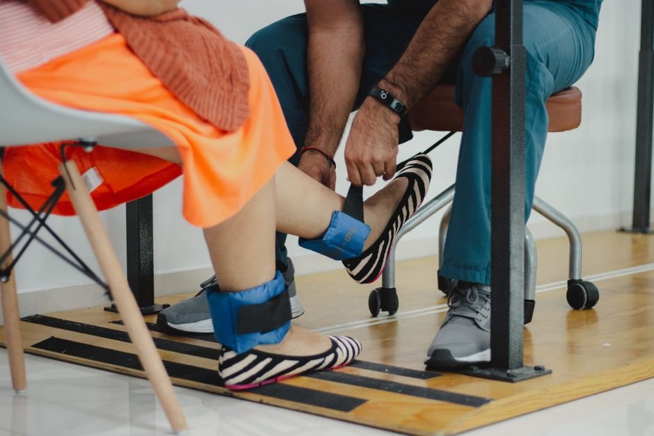 A child stepping along a low balance beam in a bright therapy room while a physical therapist walks alongside.