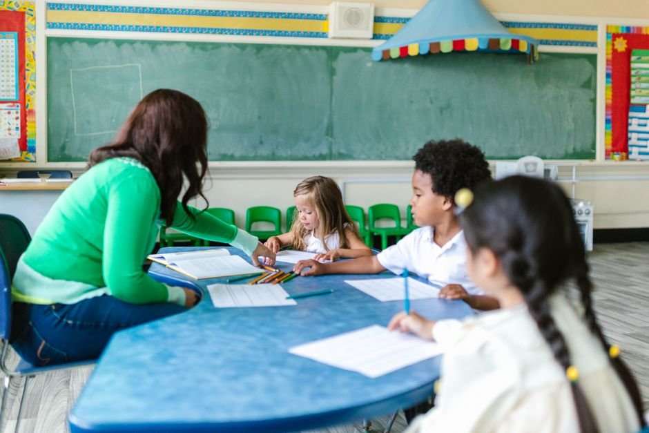 A teacher kneels beside a student's desk in a classroom while other children work at nearby tables.
