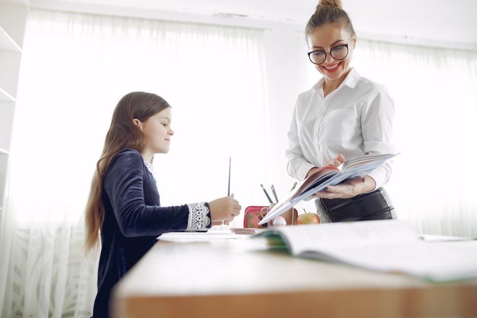 A classroom aide kneels beside a student's desk, pointing to a worksheet while the student writes.