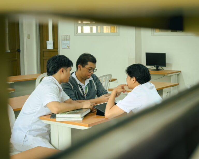A parent sits at a table with two school staff members, papers and a laptop in front of them.