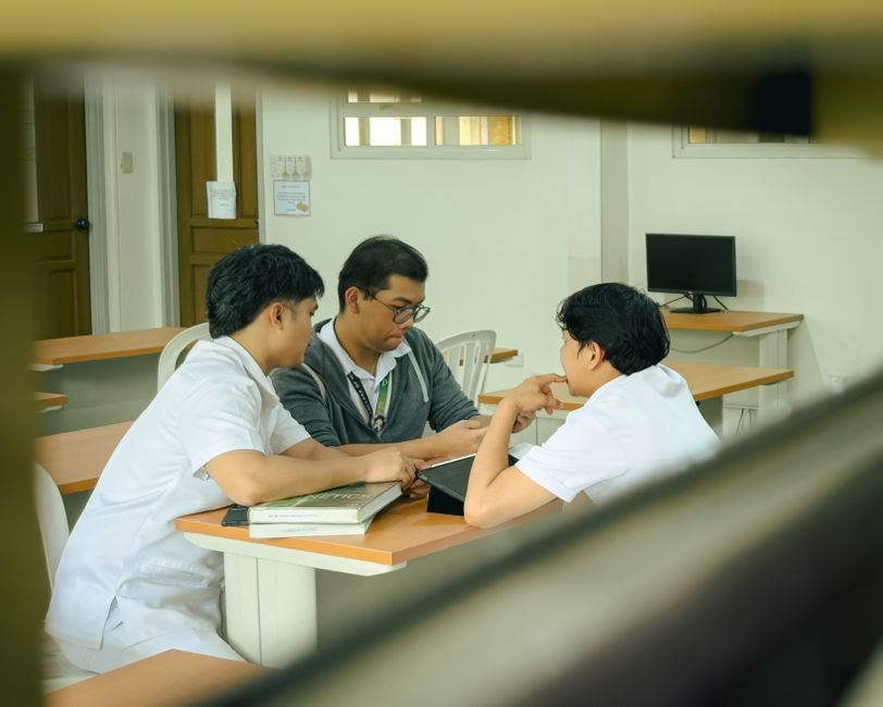 A parent sits at a table with two school staff members, papers and a laptop in front of them.