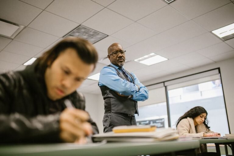 An adult with a notebook sits at the side of a classroom while students work at their desks.