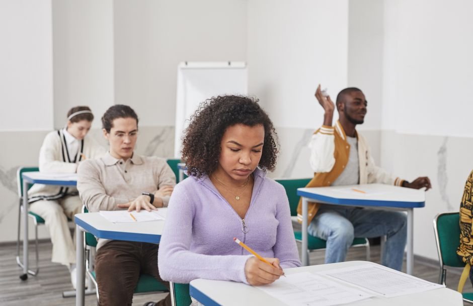 A student sits alone at a desk in a quiet, bright room, writing on a test paper.