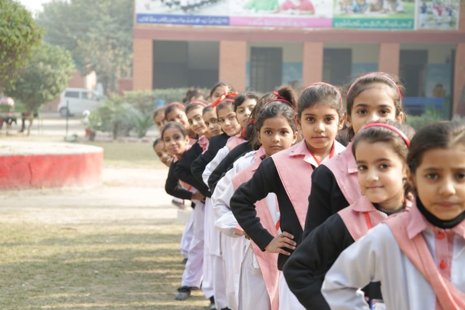 A group of students stands in a line on a school playground with a teacher beside them.