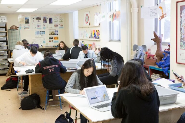Four students sit around a classroom table with markers and a large sheet of paper between them.