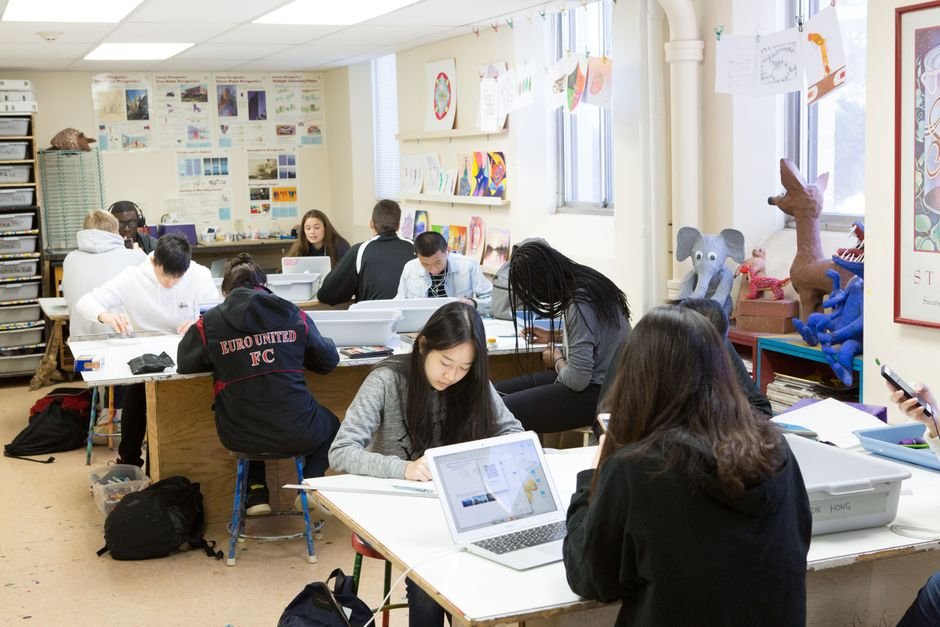 Four students sit around a classroom table with markers and a large sheet of paper between them.