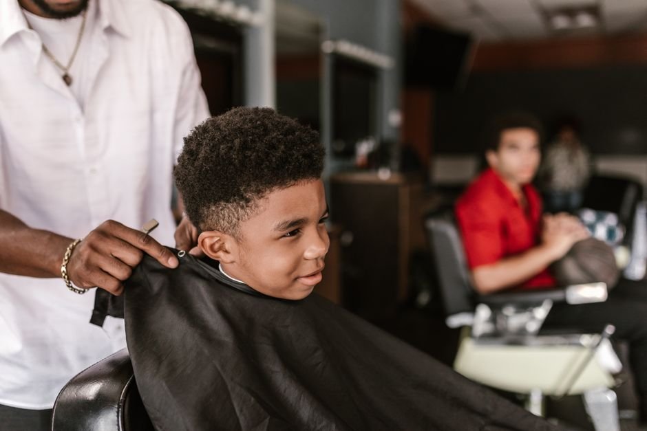 A young child sitting in a barber chair while a barber trims their hair with scissors