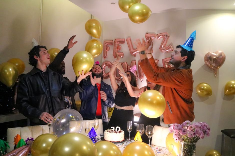 Three children sitting at a kitchen table with a small birthday cake and a few balloons